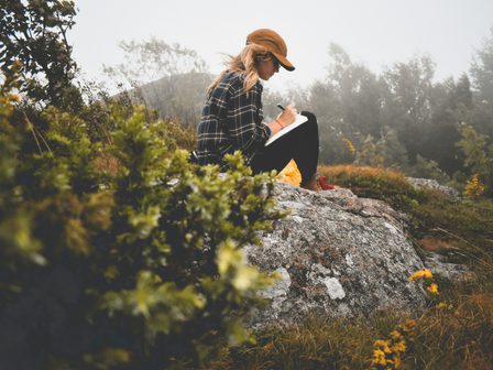 An image showing a woman sitting on a rock writing in a notepad surrounded by greenery